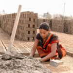A woman manually shaping bricks in an outdoor Indian factory setting.